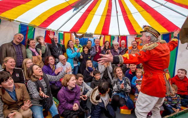 A man in circus costume performing to an audience in a colourful tent