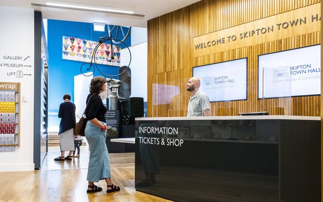 A woman talking to a man at the reception desk. The sign on the front of the desk says 'Information Tickets and Shop'.